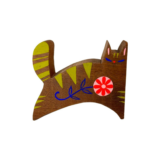 Die cut wooden sculpture of a cat, with a simplistic arched body and a fluffy tail. Painted onto the stained wood are stylistic green stripes, blue slit eyes and a red flower with a blue stem on the body. 