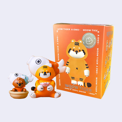 Small orange tiger figure with a large fish shaped hat around its head. It sits and holds a pink lotus. Next to it is a small brown bear in a bowl with a similar hat. They both stand in front of their blind box packaging.