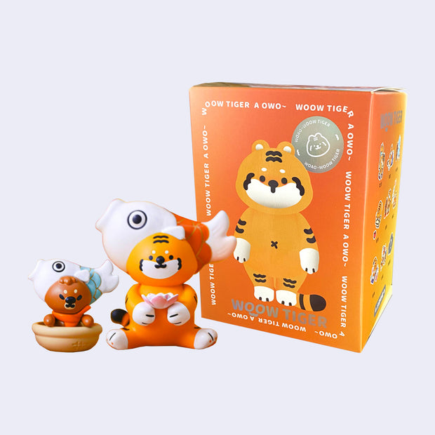 Small orange tiger figure with a large fish shaped hat around its head. It sits and holds a pink lotus. Next to it is a small brown bear in a bowl with a similar hat. They both stand in front of their blind box packaging.