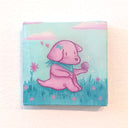 A square canvas painting featuring a pink puppy sitting in a blue grassy field under a light blue sky with pink clouds. It holds a sparkling pink ball in its paw.