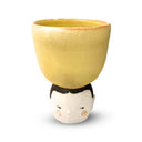 Yellow ceramic cup with the head of a person with a small, stoic expression, rosy cheeks and short black hair as the cup's "handle".