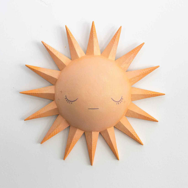 Painted pastel orange sculpture of a cartoon style sun, with a rounded dome center and 16 spikes as sun rays. It has a simple closed eye face with a straight lined mouth and no nose or other facial features.