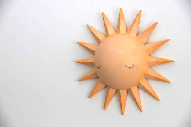 Painted pastel orange sculpture of a cartoon style sun, with a rounded dome center and 16 spikes as sun rays. It has a simple closed eye face with a straight lined mouth and no nose or other facial features. Shown at a side angle.