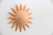 Painted pastel orange sculpture of a cartoon style sun, with a rounded dome center and 16 spikes as sun rays. It has a simple closed eye face with a straight lined mouth and no nose or other facial features. Shown at a side angle.
