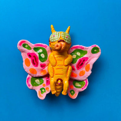 Clay figure of a yellow bodied butterfly man with pink wings with green, orange and pink designs on it. 