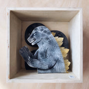 Shadowbox-esque sculpture of Godzilla, colored gray with watercolor style pattern and gold spikes on its back. Only its upper body is showing.