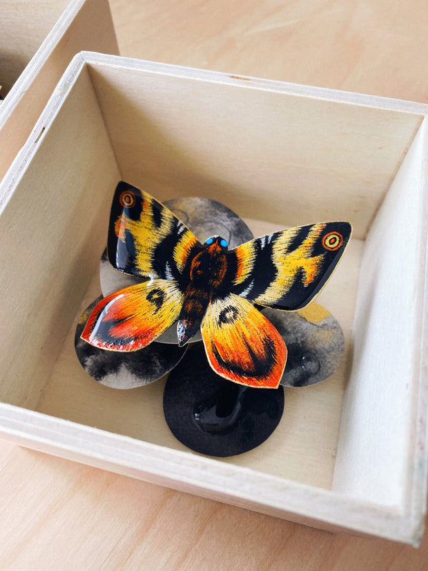 A shadow-box-esque sculpture containing an illustration of Mothra, posited on grayish black circles.