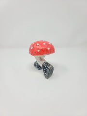 Back angle of small sculpture of a red mushroom with white dots, with legs in pinstriped pants growing out of the mushroom cap, in mid-step position. It wears black and white polka dot sneakers. It looks as if it's running away.