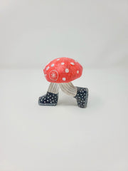 Side angle of small sculpture of a red mushroom with white dots. It has a cartoon smiling face, with legs in pinstriped pants growing out of the mushroom cap, in mid-step position. It wears black and white polka dot sneakers.