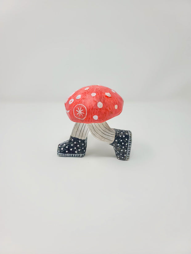 Side angle of small sculpture of a red mushroom with white dots. It has a cartoon smiling face, with legs in pinstriped pants growing out of the mushroom cap, in mid-step position. It wears black and white polka dot sneakers.