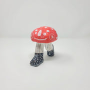 Small sculpture of a red mushroom with white dots. It has a cartoon smiling face, with legs in pinstriped pants growing out of the mushroom cap, in mid-step position. It wears black and white polka dot sneakers.