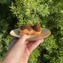 VIdeo of someone rotating a sculpture of a sleeping tan cat, with simplistic facial features and a red smile. Cat has darker brown accent coloring towards the top of its body, with green flecks on its back to make it resemble a takoyaki.