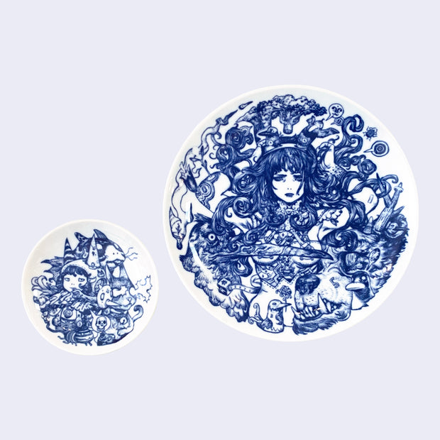 Set of two white ceramic plates with deep blue intricate line art illustrations. A small plate features an illustration of a girl surrounded by various animals and animal masks. Larger plate features a woman with lots of dark, wavy hair, surrounded by a large bird with a long beak and a grizzled bear with an underbite.
