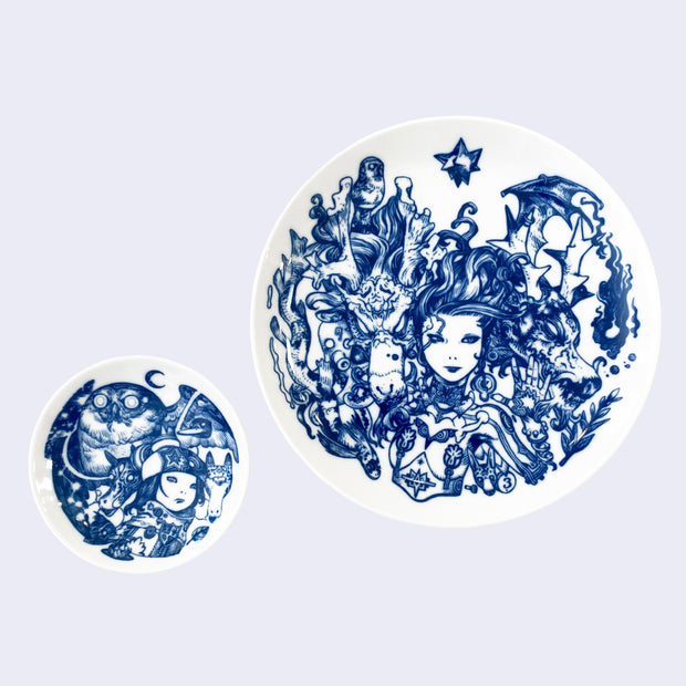 Two white ceramic plates with intricate blue line art illustrations. Smaller plate features a girl with a large owl behind her and two horse-like heads at her side. Larger plate features a woman with long, wavy hair and mechanical body, one hand lightly touching a dragon head, with another dragon is on her opposite side.
