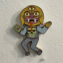 Illustrated wood cut of yellow goblin with three eyes, wearing a sweater vest with a skull. It wears a hand mitt designed as a monster.