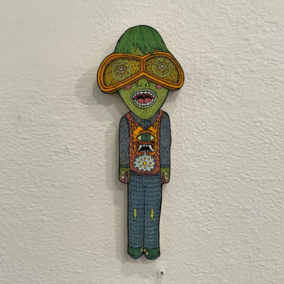 Illustrated wood cut of a green shaggy haired humanoid with large patterned goggles. It stands straight with a detailed patterned outfit and orange monster on his sweater vest.