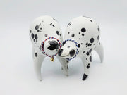 Rounded white ceramic sculpture of a quadruped with black polka dots. It has an mouth and a woven red, white and blue collar with a gold bell on it. It has a wide, open mouth smile. It stands next to a twin sculpture, that has a blue and white collar.