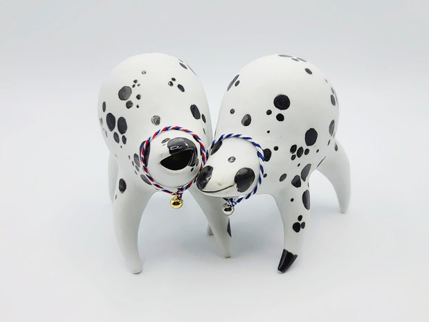 Rounded white ceramic sculpture of a quadruped with black polka dots. It has a woven white and blue collar with a gold bell on it. It has a wide, slightly open mouth smile. It stands next to a nearly identical sculpture, with a red, white and blue collar.