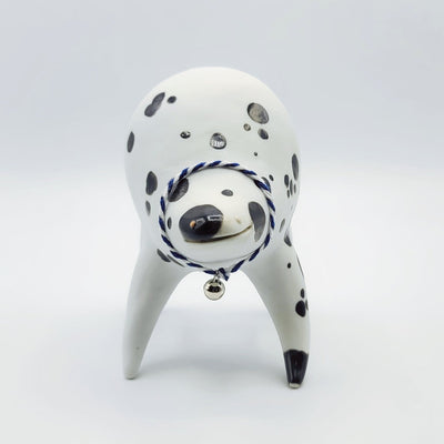Rounded white ceramic sculpture of a quadruped with black polka dots. It has a woven white and blue collar with a gold bell on it. It has a wide, slightly open mouth smile.