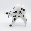 Rounded white ceramic sculpture of a quadruped with black polka dots. It has a woven white and blue collar with a gold bell on it. It has a wide, slightly open mouth smile.