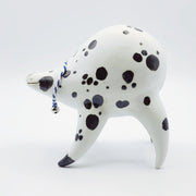 Rounded white ceramic sculpture of a quadruped with black polka dots. It has a woven white and blue collar with a gold bell on it. It has a wide, slightly open mouth smile.