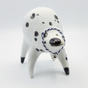 Rounded white ceramic sculpture of a quadruped with black polka dots. It has a woven white and blue collar with a gold bell on it. It has a wide, slightly open mouth smile.