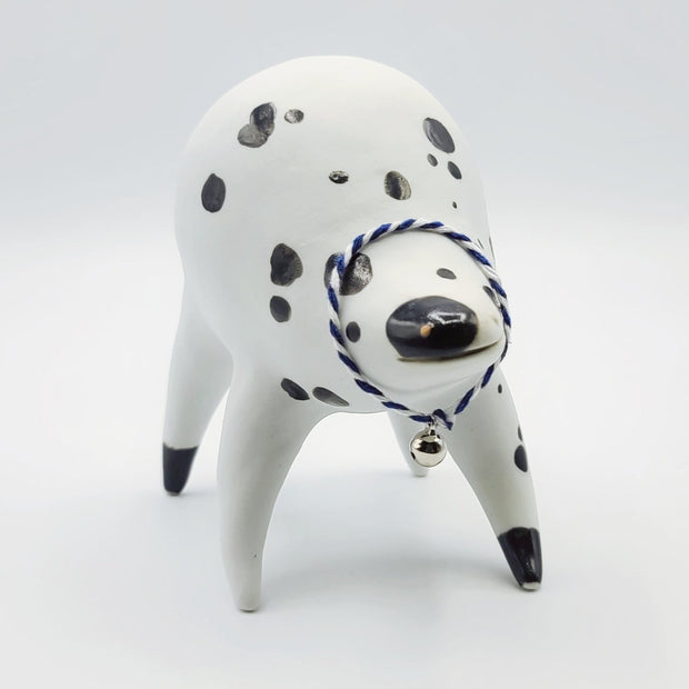 Rounded white ceramic sculpture of a quadruped with black polka dots. It has a woven white and blue collar with a gold bell on it. It has a wide, slightly open mouth smile.