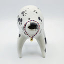 Rounded white ceramic sculpture of a quadruped with black polka dots. It has a woven red, white and blue collar with a gold bell on it. It has a wide, open mouth smile.