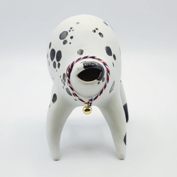 Rounded white ceramic sculpture of a quadruped with black polka dots. It has a woven red, white and blue collar with a gold bell on it. It has a wide, open mouth smile.