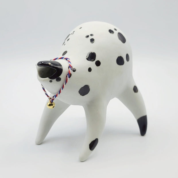 Rounded white ceramic sculpture of a quadruped with black polka dots. It has a woven red, white and blue collar with a gold bell on it. It has a wide, open mouth smile.