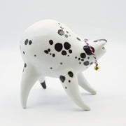 Rounded white ceramic sculpture of a quadruped with black polka dots. It has an mouth and a woven red, white and blue collar with a gold bell on it. It has a wide, open mouth smile.