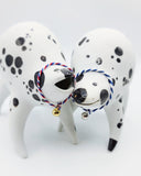 Rounded white ceramic sculpture of a quadruped with black polka dots. It has a woven red, white and blue collar with a gold bell on it. It has a wide, open mouth smile. It stands next to a nearly identical sculpture, with a blue and white collar.