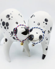 Rounded white ceramic sculpture of a quadruped with black polka dots. It has a woven red, white and blue collar with a gold bell on it. It has a wide, open mouth smile. It stands next to a nearly identical sculpture, with a blue and white collar.