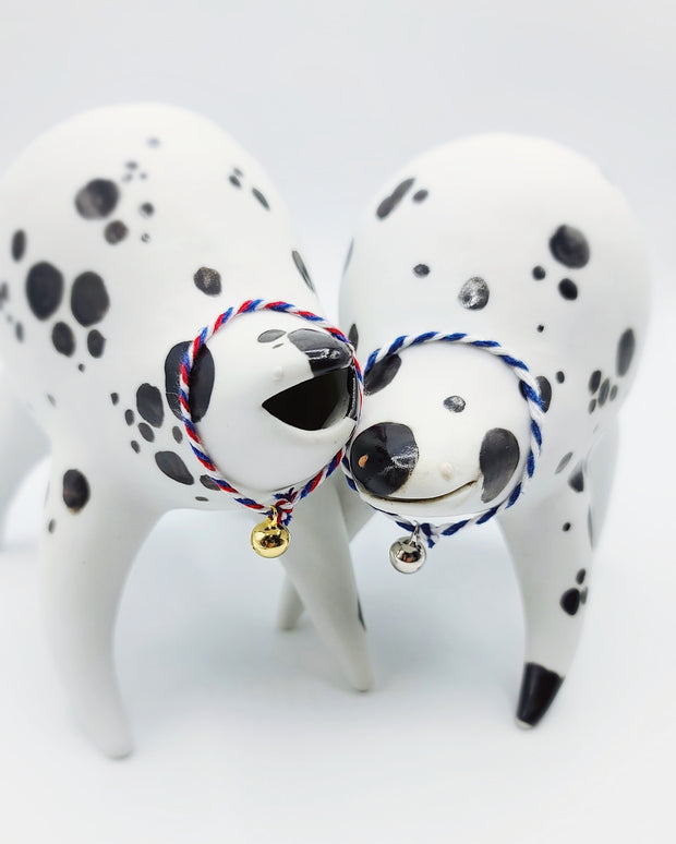 Rounded white ceramic sculpture of a quadruped with black polka dots. It has a woven white and blue collar with a gold bell on it. It has a wide, slightly open mouth smile. It stands next to a nearly identical sculpture, with a red, white and blue collar.