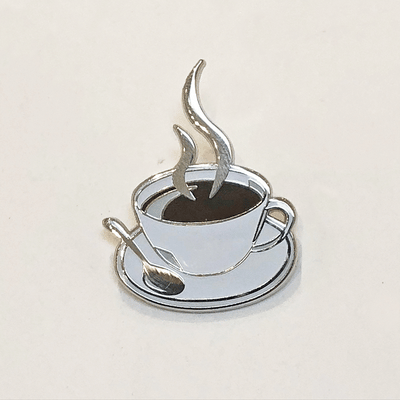 Gif of enamel pin of a cup of black coffee, in a shallow white mug on a saucer with a spoon. Illustrated steam is coming up from the coffee. One image is of the pin in the light, the other is of the pin glowing in the dark.