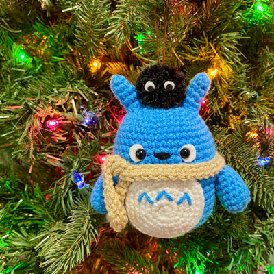 Crocheted blue chibi Totoro, wearing a cream colored scarf with a dust sprite on his head. Totoro is strung onto a Christmas tree.