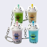 4 different designed keychains of clear to go drink cups with either green, yellow, purple or blue liquid with boba. Each have whipped cream at the top shaped like a smiling kawaii cat and a straw that matches the liquid color.