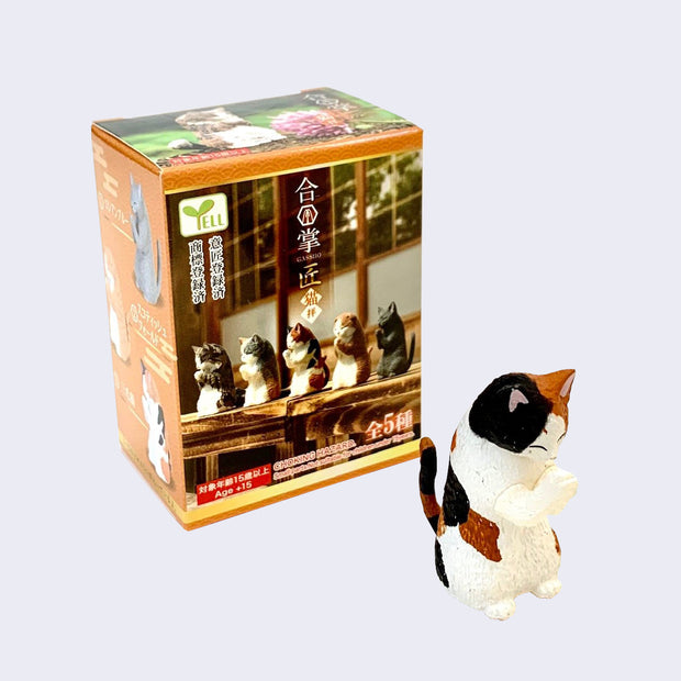 Small plastic figurine of a calico cat, standing on its hind legs and slightly hunched over as though it's bowing or praying. It stands next to its product packaging.