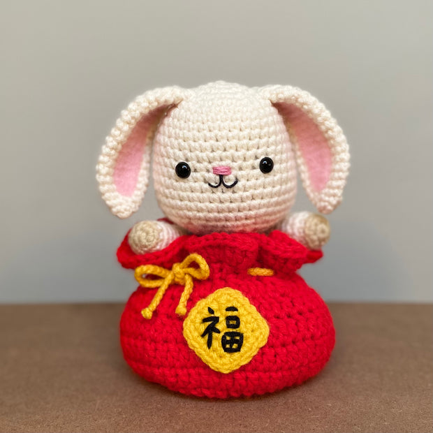 Crochet plush sculpture of a cute looking white rabbit with simple features and a smile, sitting within a red pouch that is tied up with a gold string and has Chinese script on a gold patch on the center of the pouch.