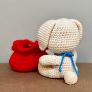 Crochet plush sculpture of a cute looking white rabbit with simple features and a smile, wearing a blue string with a bell on it. It is sitting next to a red pouch that is tied up with a gold string and has Chinese script on a gold patch on the center of the pouch.