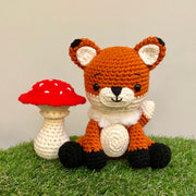 Crocheted sculpture of a cute looking fox, orange with white details and black feet and ear tips, sitting next to a crocheted red toadstool mushroom with white dots.