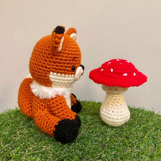 Crocheted sculpture of a cute looking fox, orange with white details and black feet and ear tips, sitting next to a crocheted red toadstool mushroom with white dots.