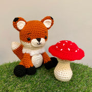 Crocheted sculpture of a cute looking fox, orange with white details and black feet and ear tips, sitting next to a crocheted red toadstool mushroom with white dots.