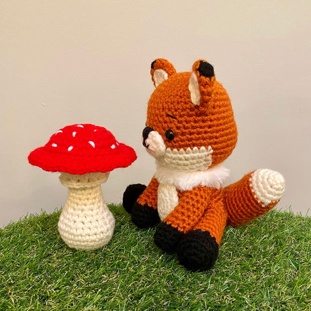 Crocheted sculpture of a cute looking fox, orange with white details and black feet and ear tips, sitting next to a crocheted red toadstool mushroom with white dots.