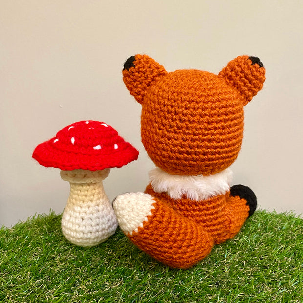 Crocheted sculpture of a cute looking fox, orange with white details and black feet and ear tips, sitting next to a crocheted red toadstool mushroom with white dots.