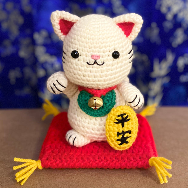 White crocheted maneki, a lucky cat with one of its paws up and a bell around its neck with a gold token near it. It sits atop of a red crocheted cushion, with gold tassels on each corner. It has black beads for eyes and a simplistic smile.