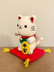 White crocheted maneki, a lucky cat with one of its paws up and a bell around its neck with a gold token near it. It sits atop of a red crocheted cushion, with gold tassels on each corner. It has black beads for eyes and a simplistic smile.