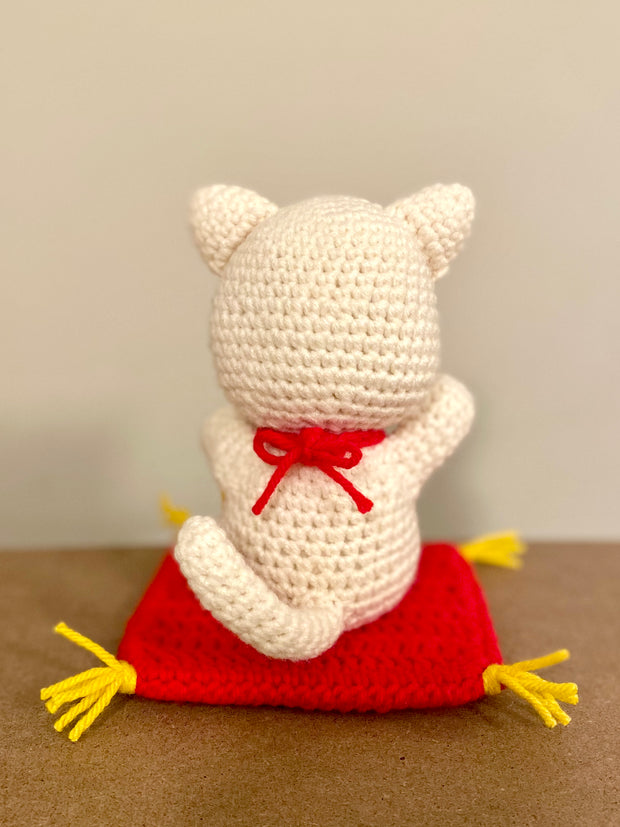 Back view of a crocheted white cat, with a mid length tail and a red bow tied around the back of its neck. It sits atop of a red crocheted cushion with gold tassels on each side.
