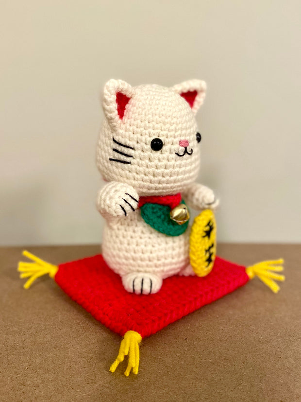 White crocheted maneki, a lucky cat with one of its paws up and a bell around its neck with a gold token near it. It sits atop of a red crocheted cushion, with gold tassels on each corner. It has black beads for eyes and a simplistic smile.