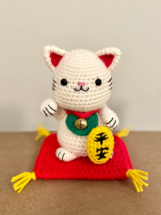 White crocheted maneki, a lucky cat with one of its paws up and a bell around its neck with a gold token near it. It sits atop of a red crocheted cushion, with gold tassels on each corner. It has black beads for eyes and a simplistic smile.